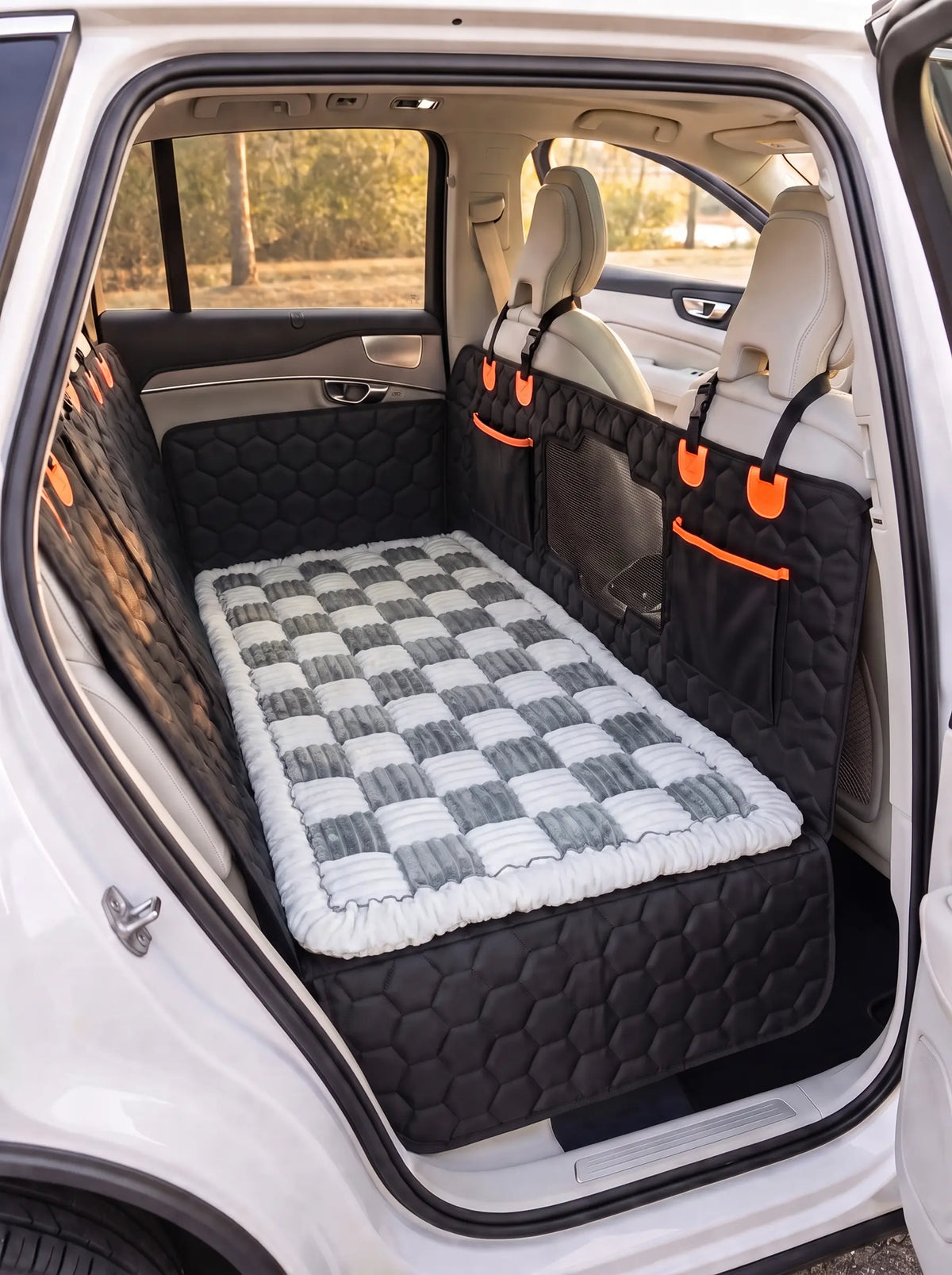 A pet car bed with a gray and white checkered pattern and a white faux fur trim, shown installed in the back of a white SUV. The bed is fitted between the rear seats on top of the silverhammock car seat cover